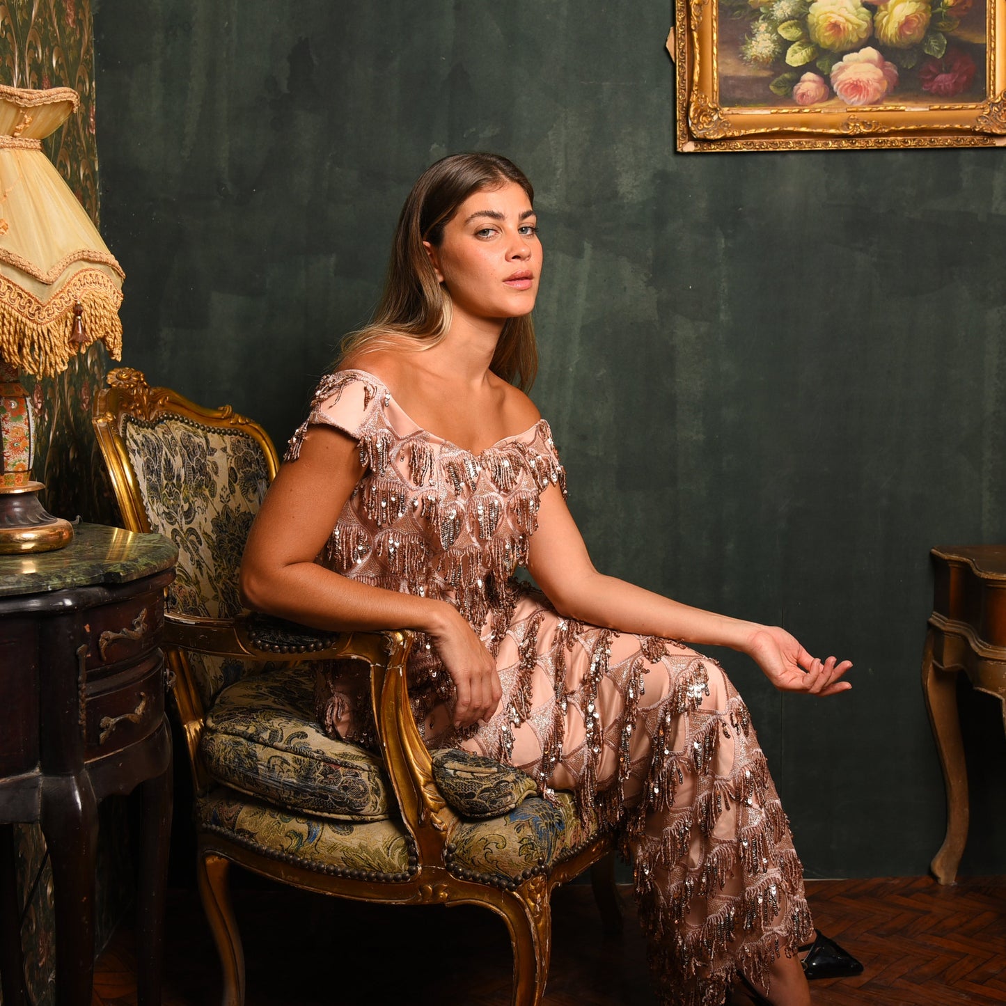 Woman in a patterned dress sitting on an ornate chair in a vintage room with a lamp and painting